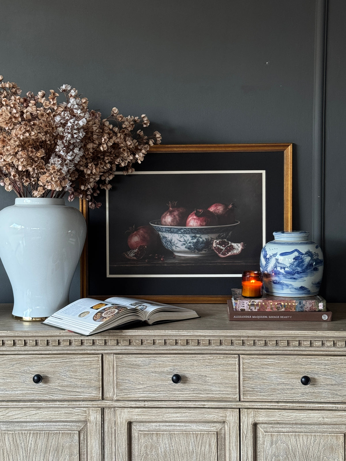 Willow Mist Landscape Jar on a stack of books beside a candle and a still life of pomegranates