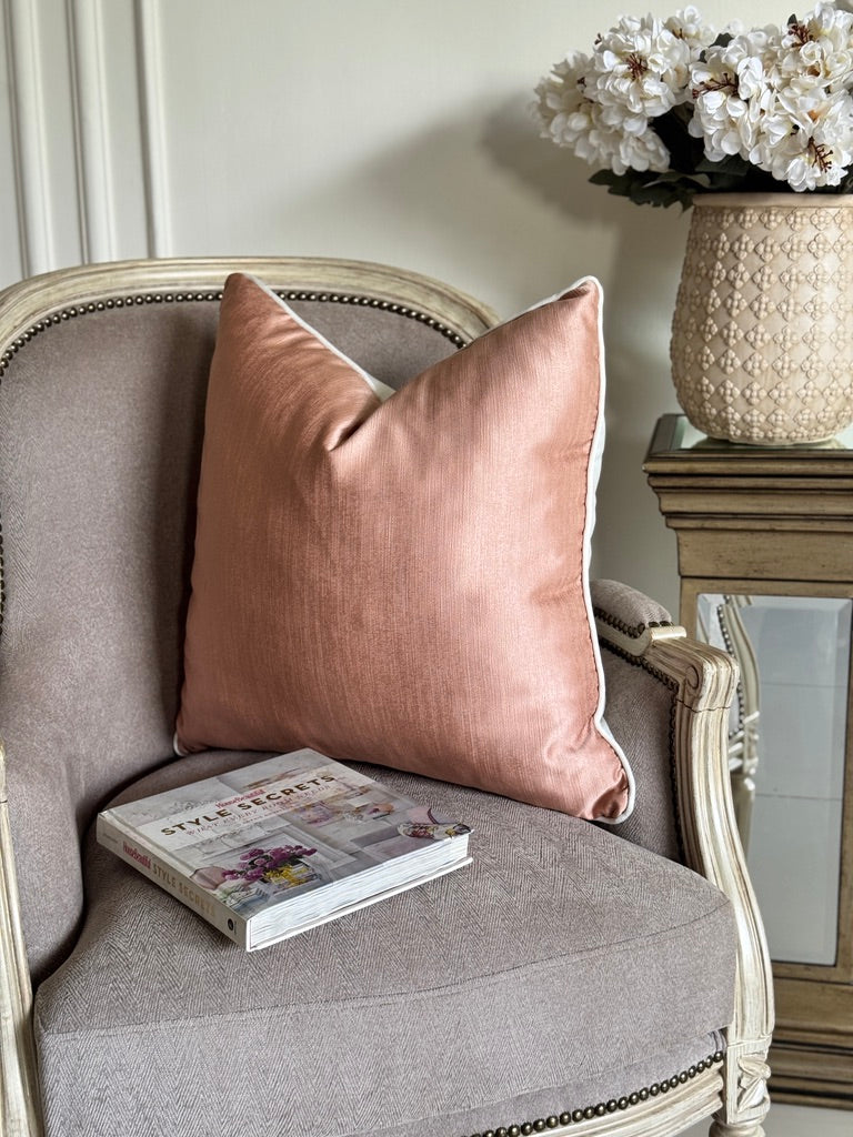 A dusty rose velvet throw pillow with white piping rests on a gray upholstered armchair, with a book and a vase of white flowers in the background.