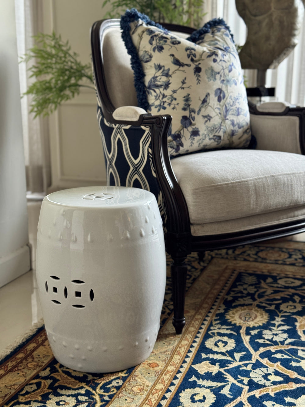 A white ceramic barrel-shaped side table with decorative cutout details, styled next to a classic armchair with blue and white patterned cushions on a patterned rug.