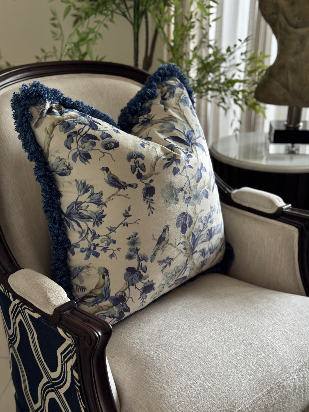 A decorative throw pillow with a blue floral and bird toile pattern on a cream background, featuring dark blue fringed edges, styled on a beige upholstered armchair in a home setting.