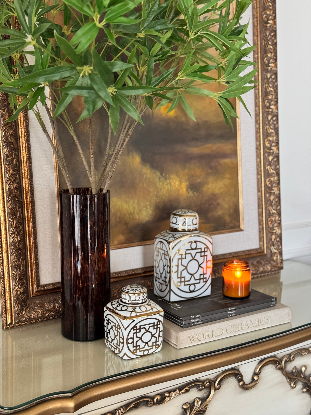 Close-up of faux bamboo leaf stems in a tortoiseshell vase with white and gold lattice carafes and an amber candle on a console – Bohemia Lifestyle