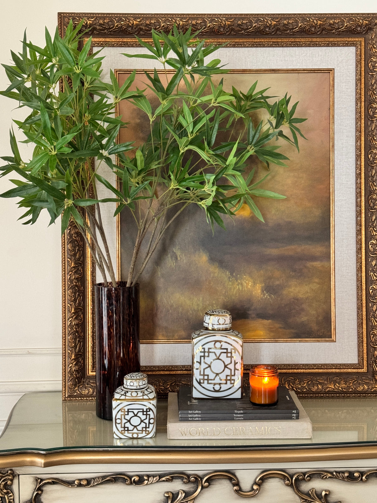 A styled interior scene with a large ornate gold-framed landscape painting leaning against a wall. In front of the frame, a tall dark vase holds green bamboo stems, next to two decorative white ceramic canisters and a small lit orange candle on stacked books.